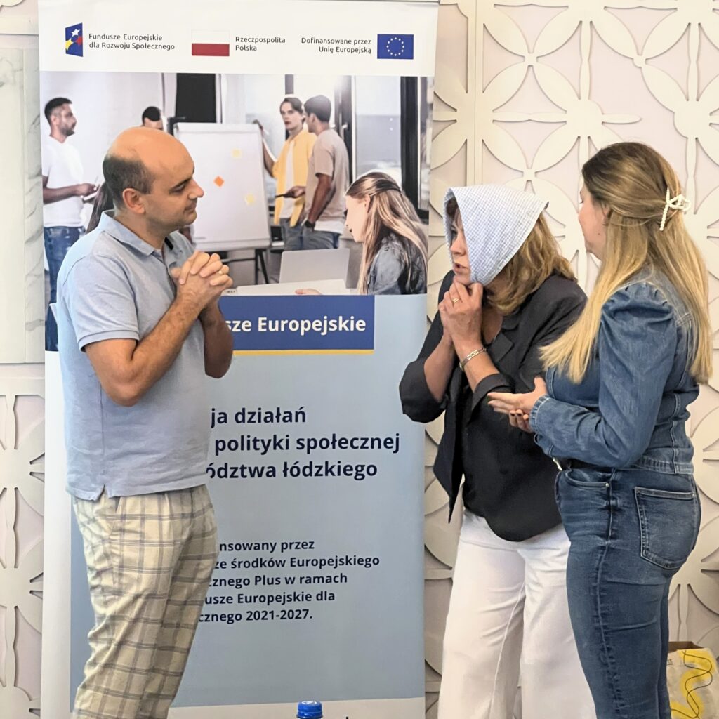 3. Trzy osoby dorosłe stoją przed plakatem konferencyjnym. Mężczyzna ma krótkie ciemne włosy, jest ubrany w niebieską koszulkę polo i spodnie od piżamy i patrzy w prawo z lekkim uśmiechem. Kobieta w środku ma blond włosy do ramion, ciemną marynarkę i kraciaste nakrycie głowy. Gestykuluje lewą ręką i wygląda na zszokowaną. Po jej prawej stronie, młodsza kobieta z długimi blond włosami w dżinsowej kurtce słucha z założonymi rękami. Plakat częściowo widoczny za nimi zawiera tekst w języku polskim oraz logo Unii Europejskiej i Funduszy Europejskich dla Rozwoju Społecznego.
4. Grupa składająca się z jedenastu osób siedzi przy długim, prostokątnym stole