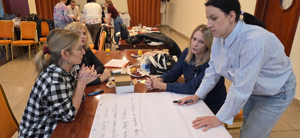 Grupa uczestniczek siedzi przy stole i pracuje nad dużą kartą papieru z notatkami; jedna osoba stoi i zapisuje wnioski. Sala szkoleniowa, na stołach materiały i napoje.
