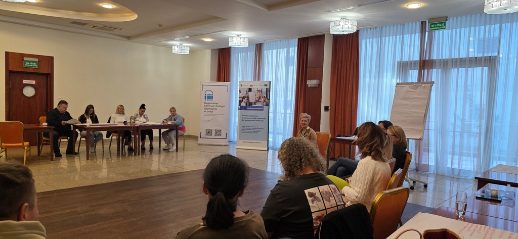 Uczestnicy warsztatów siedzą w półokręgu w sali konferencyjnej; na tle roll-upy projektu i tablica. Prowadząca moderuje dyskusję.