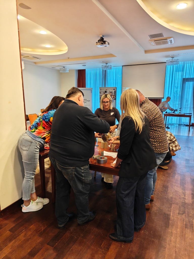 Niewielka grupa stoi wokół stołu i buduje model z papieru, tekturowych rurek i taśmy. Praca manualna w zespole.