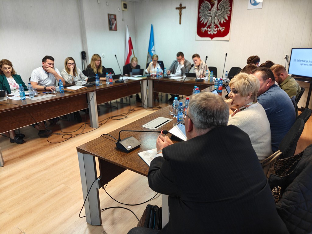 posiedzenie w sali rady — uczestnicy przy stołach w kształcie litery U, laptopy i mikrofony.