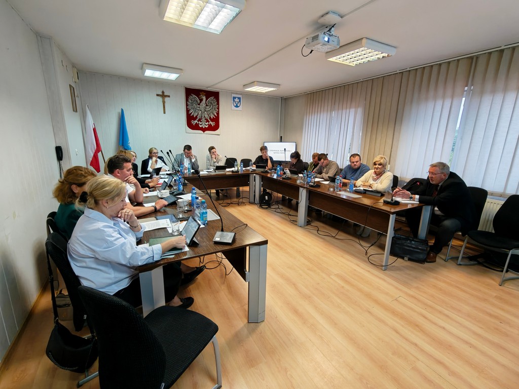 Posiedzenie w sali rady — uczestnicy przy stołach w kształcie litery U, laptopy i mikrofony.