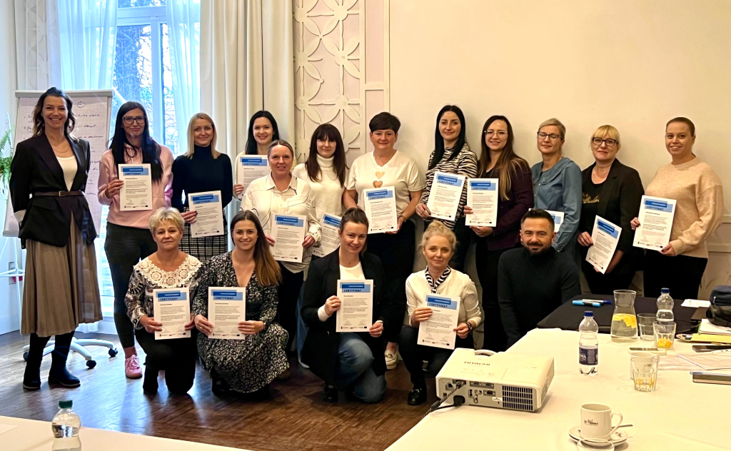 1 Grupa osób uczestniczących w szkoleniu CUS stoi w sali szkoleniowej, trzymając w rękach certyfikaty. Osoby stają na tle ściany z oknem, widać jasne zasłony, lampę.