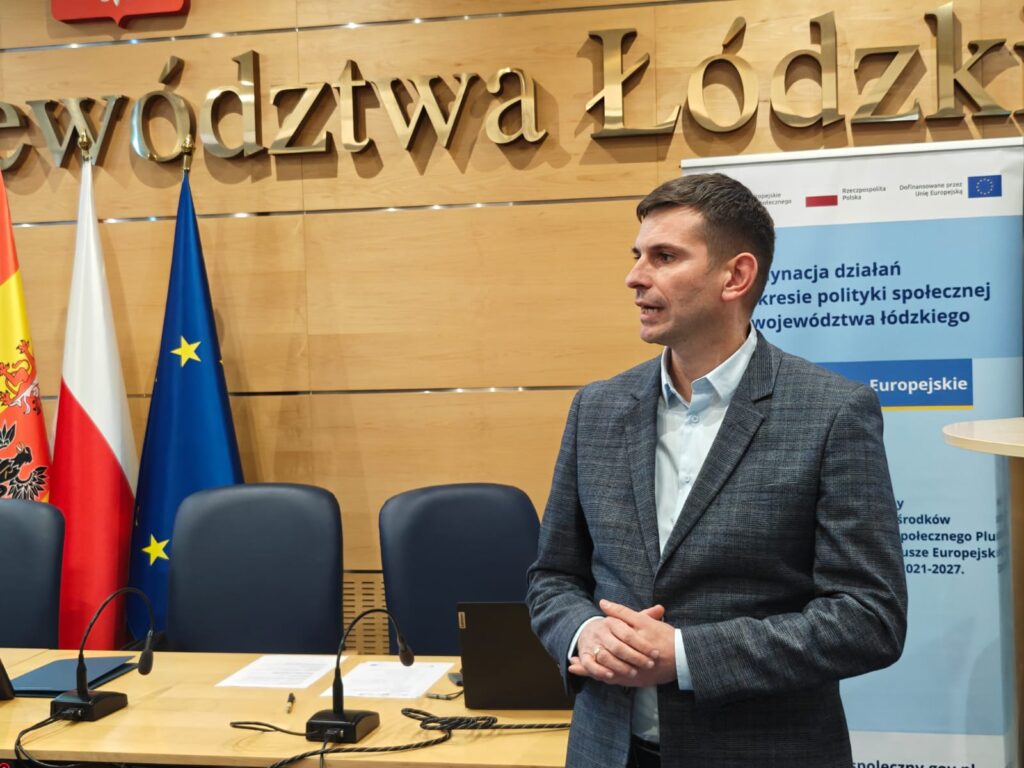 2.	Mężczyzna w garniturze prezentuje się przed stołem z flagami