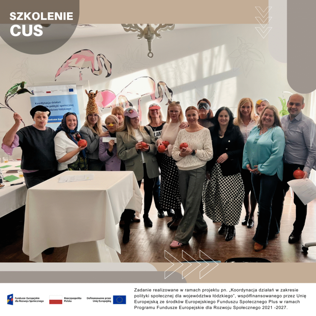 Grupa osób stoi w sali i pozuje z kolorowymi rekwizytami oraz owocami. W tle widoczne są dekoracje ścienne i jasne oświetlenie