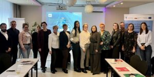 1. Grupa osób pozująca do zdjęcia przed stołem w sali szkoleniowej, w tle roll-up, ekran i flipchart.