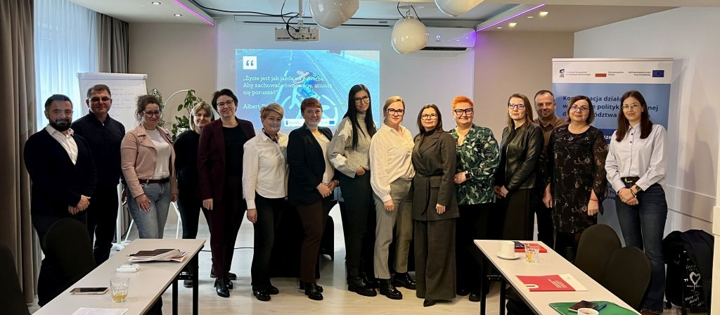 1. Grupa osób pozująca do zdjęcia przed stołem w sali szkoleniowej, w tle roll-up, ekran i flipchart.