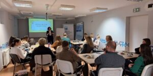 Osoby siedzą przy stołach w sali szkoleniowej i słuchają prowadzącej stojącej przed ekranem z prezentacją dotyczącą współpracy instytucji