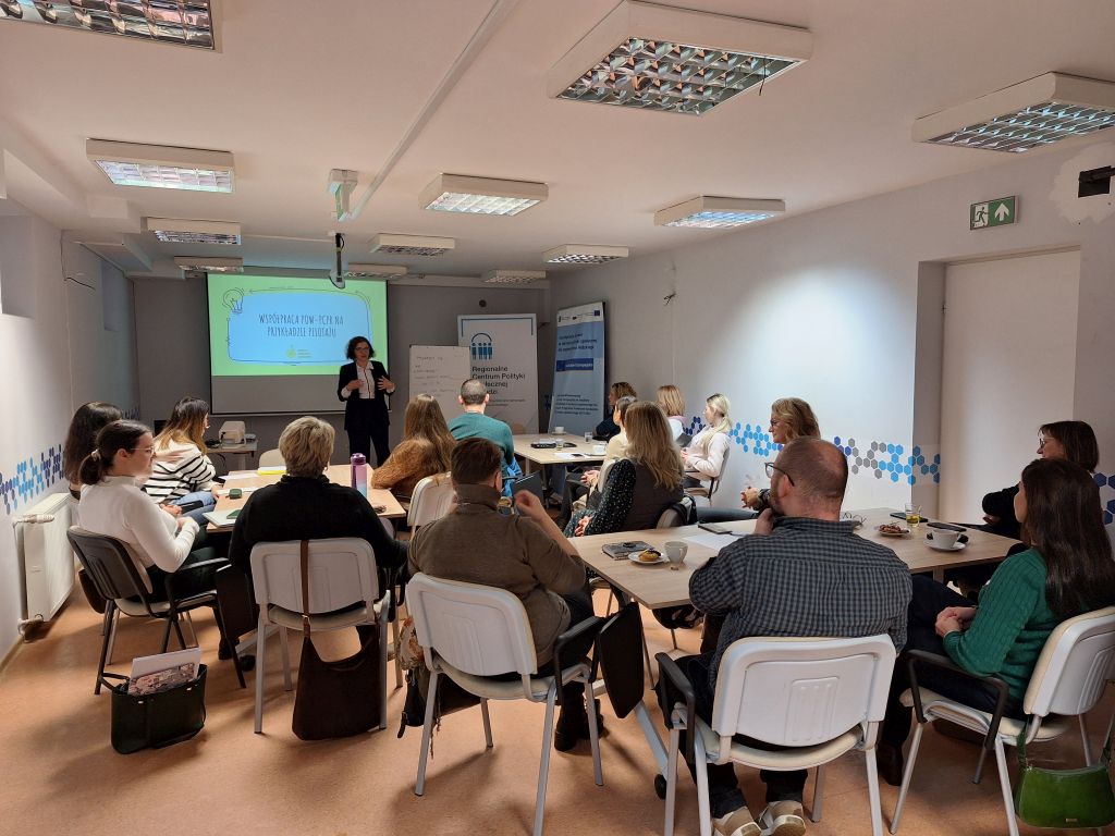 Sala szkoleniowa, w której kilkanaście osób siedzi przy stołach i słucha prowadzącej stojącej przy ekranie z prezentacją.