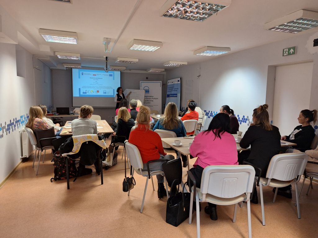 Sala szkoleniowa z kilkunastoma osobami siedzącymi przy stołach, skierowanymi w stronę prowadzącej prezentację wyświetlaną na ekranie projektora.