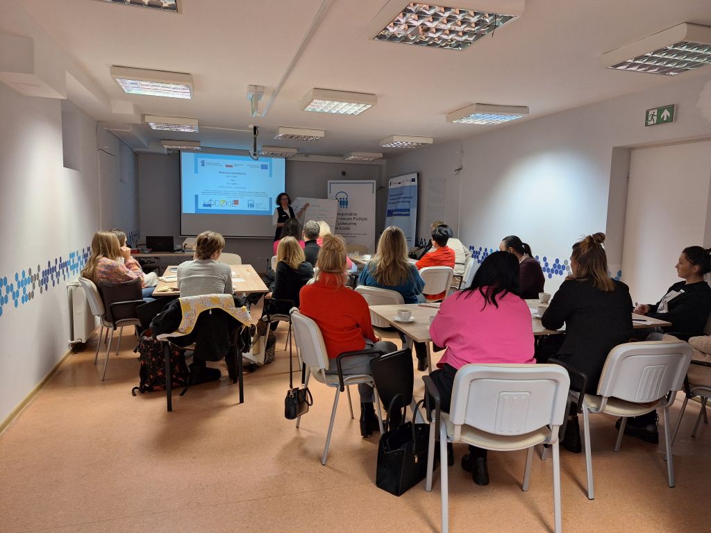 Osoby siedzą przy stołach w sali szkoleniowej i słuchają prowadzącej stojącej przed ekranem z prezentacją dotyczącą współpracy instytucji