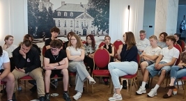 Grupa ludzi siedzących na krzesłach w sali, uczestniczących w spotkaniu lub dyskusji