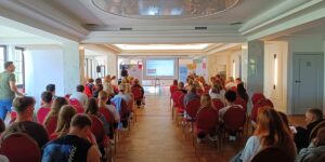 Grupa ludzi siedzących na krzesłach w sali, uczestniczących w spotkaniu lub wydarzeniu