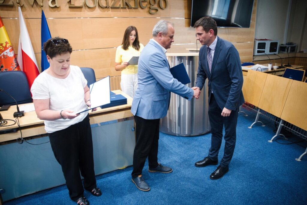 Kobieta trzyma teczkę, dwaj mężczyźni ściskają dłonie przed stołem, symbolizując do składu gremium