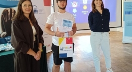 Trzy osoby stoją przed ekranem, mężczyzna trzyma certyfikat, uśmiechając się do kamery